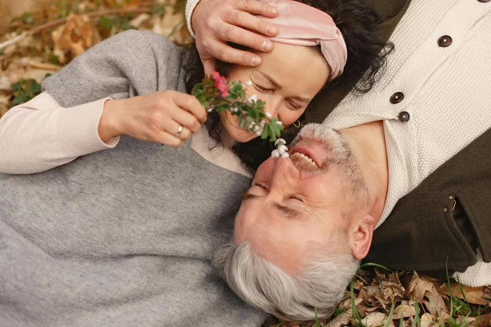 Older couple lying in autumn leaves, laughing together as she holds a sprig of flowers